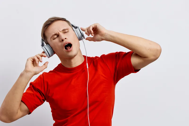 Young man wearing casual red t-shirt enjoying music with over-ear headphones, eyes closed, expressing joy and relaxation, isolated on plain white background, studio portrait. Music lifestyle concept, 