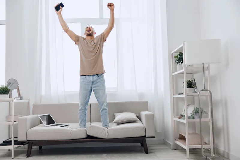 Excited man celebrating success in a bright, modern living room. He stands on the couch, raising his arms with a smartphone in hand, surrounded by minimalistic decor. ###