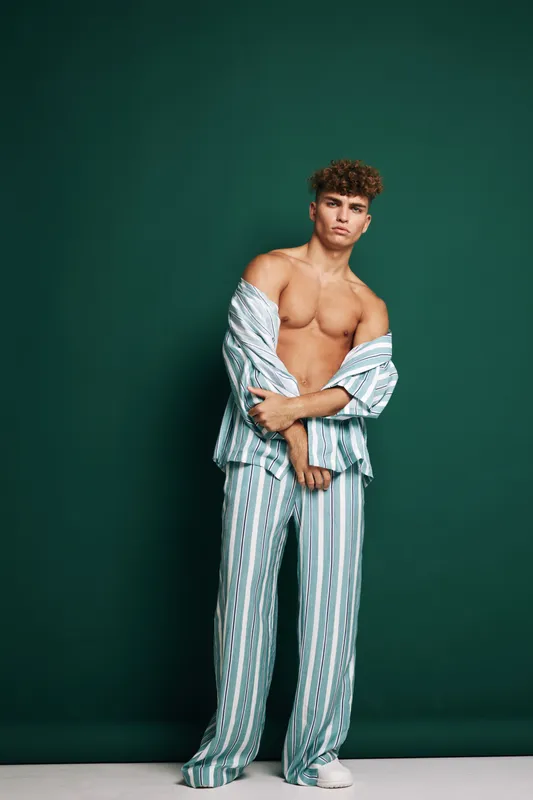 Young man with curly hair wearing striped pajamas standing against dark green background with shirt open showing bare chest and relaxed pose in studio setting.