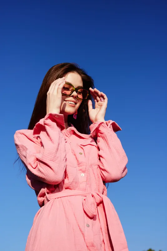 Cheerful young woman in a stylish pink dress and sunglasses, enjoying a sunny day against a bright blue sky Perfect for fashion and summer themes