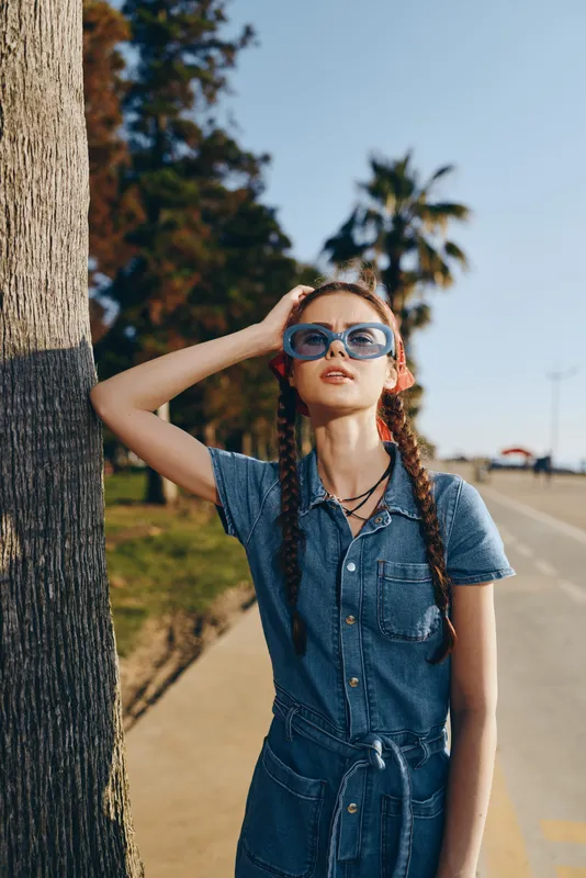 young woman in denim dress wearing blue sunglasses standing by tree near road on sunny day