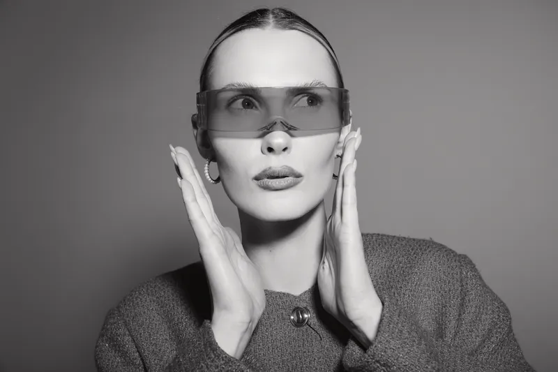 A young white woman with pale skin and slicked back hair wears large transparent futuristic glasses, touching her face with both hands. Concept of fashion, style, and modern eyewear.