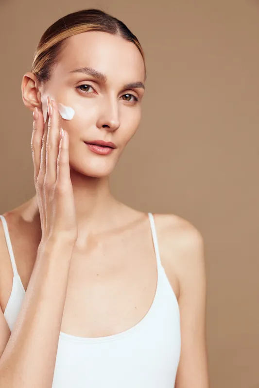 A young woman with clear skin applies white cream to her face while looking at the camera. The neutral background emphasizes skincare and natural beauty concepts.