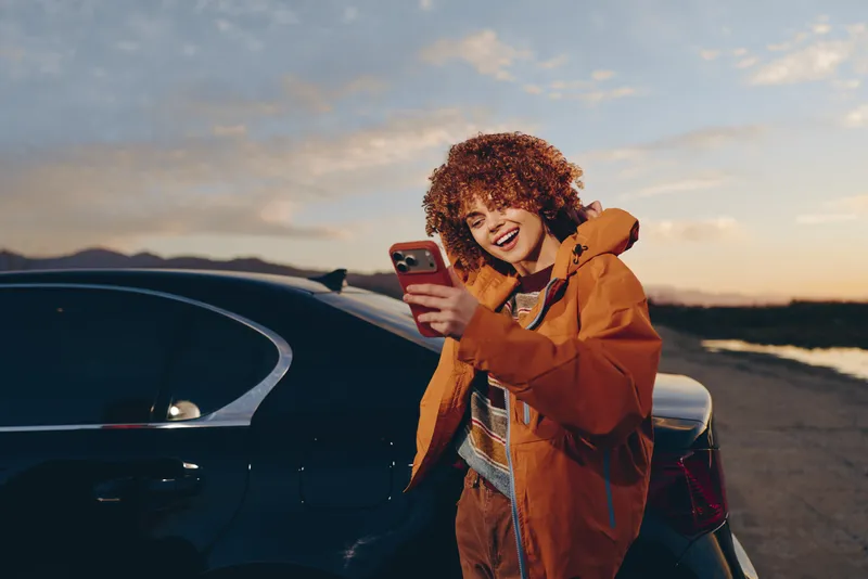 Smiling woman with curly hair in an orange jacket using a smartphone near a parked car at sunset on an open road outdoor lifestyle travel mood and warm light