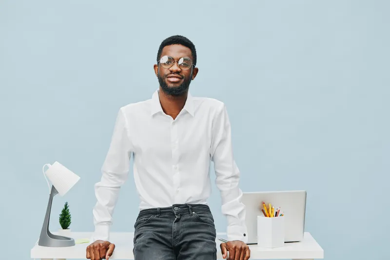 Professional portrait of a confident young African American man wearing white shirt, glasses, standing behind a modern desk with a laptop and stationery, isolated on light blue background. Business li