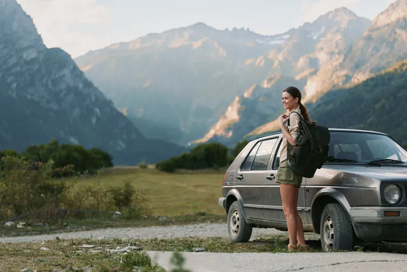 Young woman with backpack stands beside a car on a mountain road enjoying the scenic view with a smile during a sunny day outdoors.