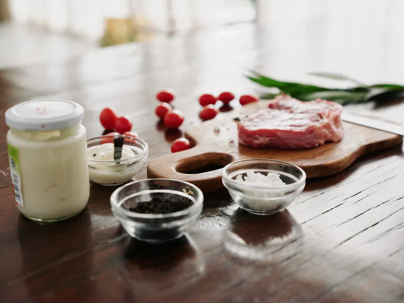 Стильная кухня: на деревянном столе лежит кусок свежего мяса со специями, помидоры и зелень, свет от окна