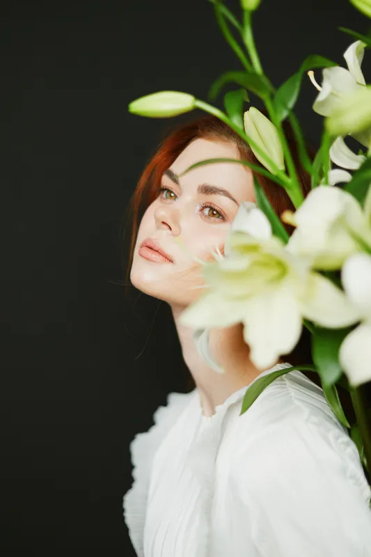Serene female model with red hair in a white dress, surrounded by white lilies against a dark background, conveying elegance and tranquility
