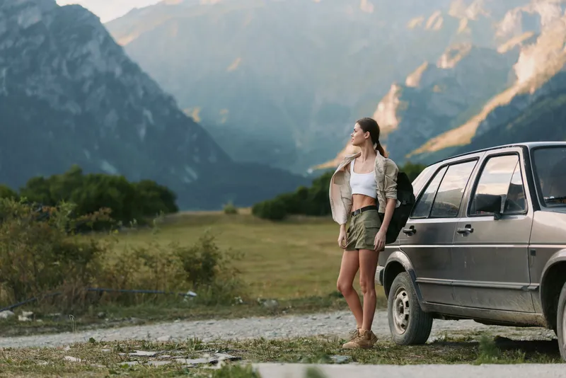 A woman in casual outdoor wear stands beside a car in a mountainous landscape, suggesting a solo travel lifestyle and outdoor exploration, with a relaxed pose, and a sense of freedom.