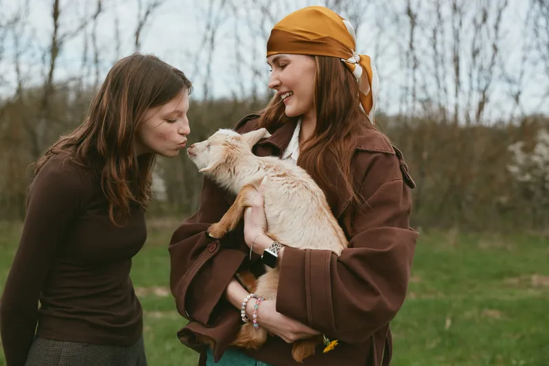 Lifestyle scene of two women outdoors with a playful goat kid, sharing smiles and gentle care, capturing a warm bond and everyday friendship in nature, casual clothing, candid moment.