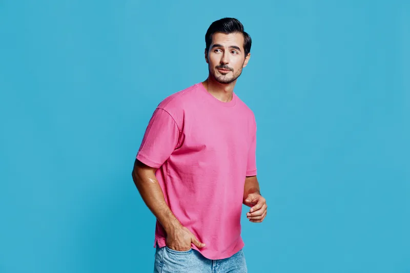 Stylish young man wearing a bright pink t-shirt stands against a vibrant blue background, looking to the side with a confident expression. Fashion lifestyle concept.