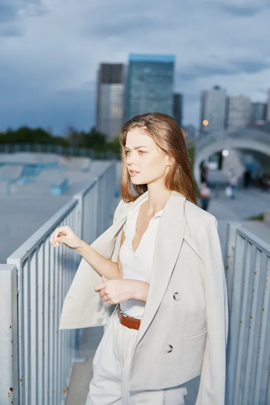 Young woman in stylish beige suit confidently walking on urban bridge during cloudy day, business fashion concept with city skyline background