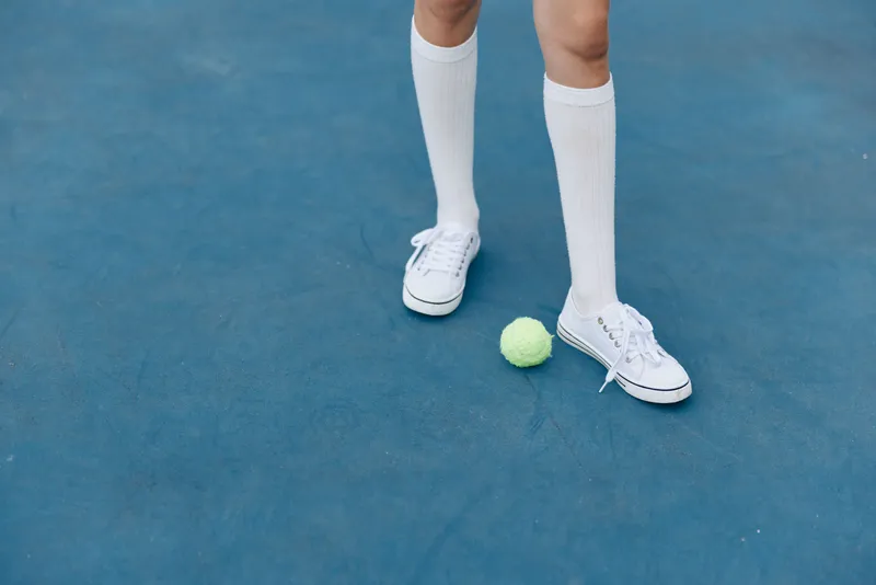 Young athlete in white knee high socks and sneakers standing on a blue tennis court with a green tennis ball, showcasing focus and determination