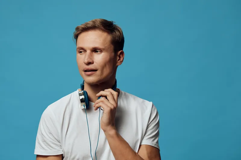 thoughtful young caucasian man with headphones hold on neck look aside wear white t-shirt isolated on blue background freelance digital nomad concept