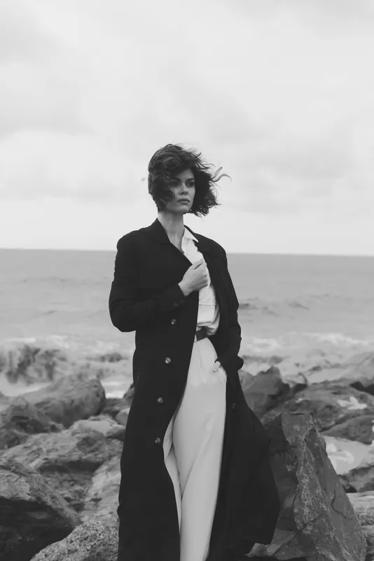 Woman on a rocky shoreline in a long coat, windblown hair and a bold stance, monochrome coastal scene conveying fashion, drama, and freedom by the sea.