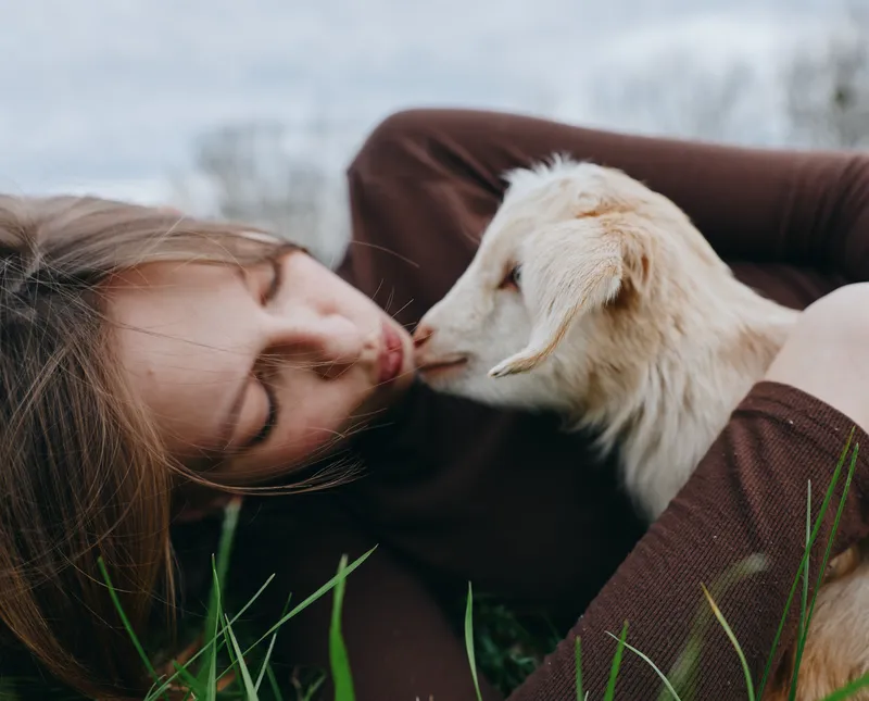 A young woman lying in the grass gently kisses a white baby goat in a natural outdoor setting, expressing tenderness and connection with the animal in a relaxed lifestyle moment.