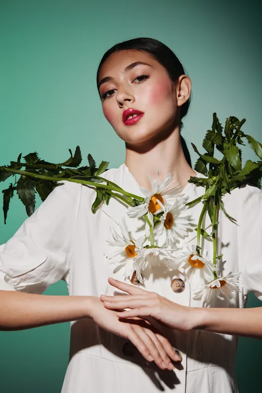 Fashion model with elaborate daisy flower arrangement and dramatic makeup poses against mint background. Creative beauty concept with unique floral decoration and stylish look.