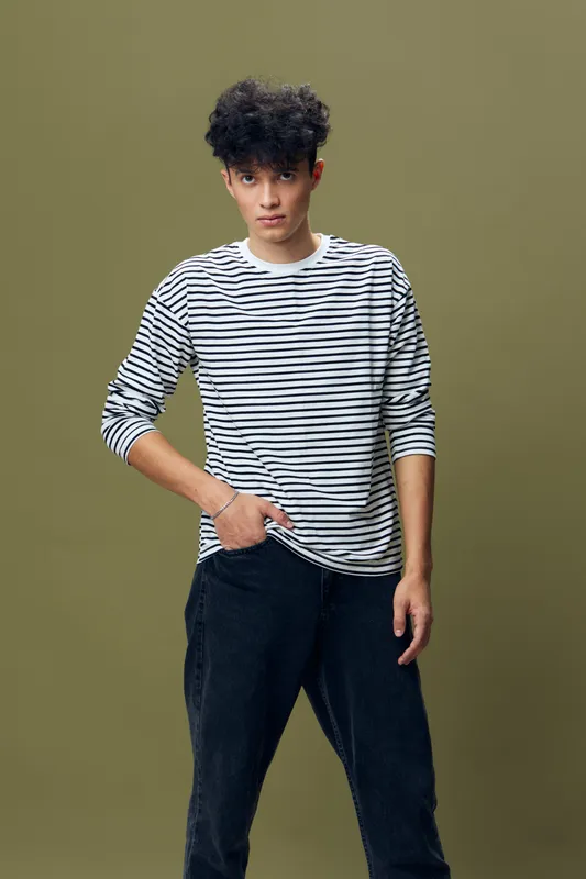 young man wearing striped shirt and black pants standing thoughtfully against olive background with casual posture and textured curly hair