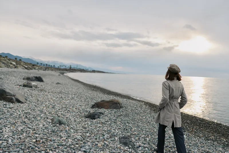 A woman in a coat and hat stands on a rocky beach looking at the sea during cloudy weather. The scene captures solitude, nature, and calmness at the coastline.