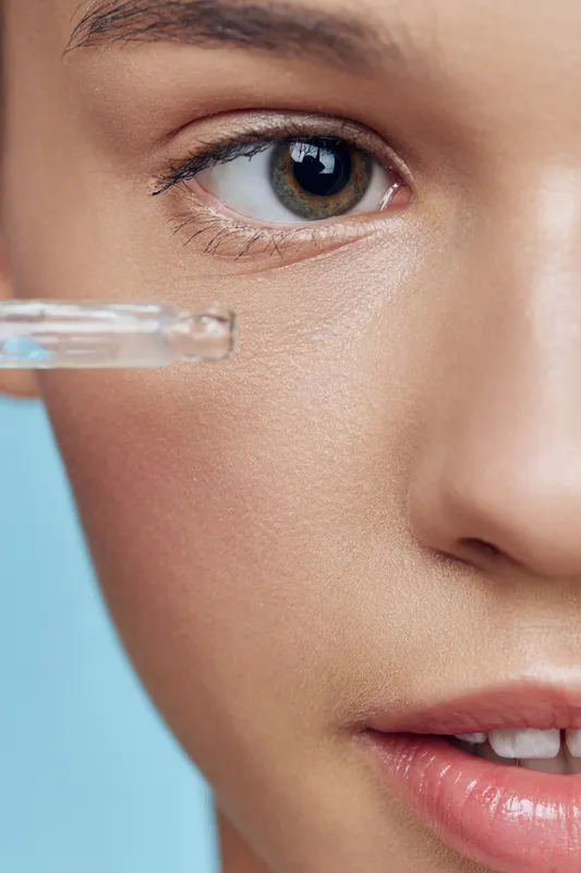 Close up of a young model with bright skin applying a skincare dropper, showcasing a fresh and healthy face with a vivid blue background