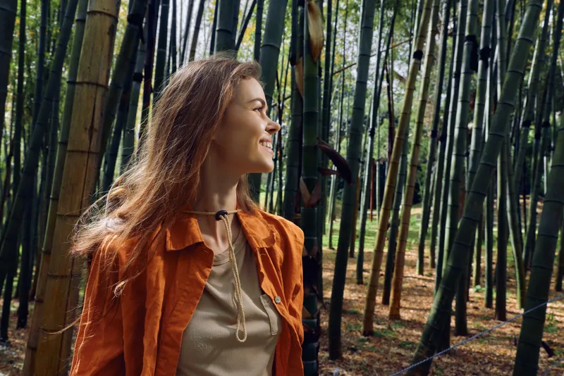 Woman bamboo forest portrait smiling and looking sideways, nature scene with tall trees and dappled sunlight, outdoors wanderer wearing orange jacket and casual clothing in woods