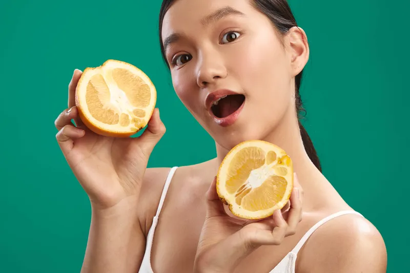 asian woman holds halved citrus in each hand against a vivid green backdrop, bright smile and surprised expression conveying freshness, energy, and playful mood.