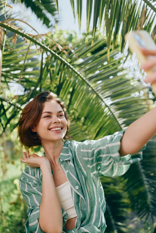 Smiling woman takes a selfie outdoors among lush palm leaves, casual green shirt, sunlit scene, bright and relaxed mood.
