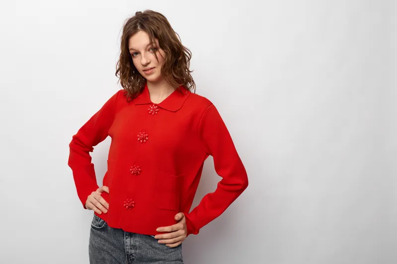 Young woman with curly hair wearing red sweater and jeans poses confidently against white background. Casual style, fashion, and relaxed mood concept for lifestyle and portrait.