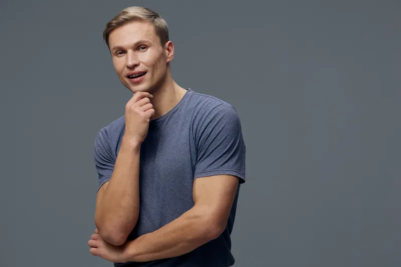 Confident smiling young man wearing casual gray t-shirt standing against plain gray background. Thoughtful expression, relaxed pose, studio portrait, lifestyle concept, cheerful mood, technology.