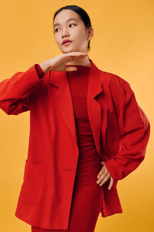 Woman in red oversized blazer and matching dress posing against yellow background with hand near chin and thoughtful expression for fashion and style concept.