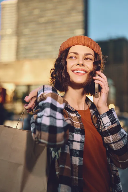 Woman shopping phone smile city beanie bag autumn scene with golden hour glow, candid lifestyle moment. Authenticity and emotional storytelling capture mindful living, joyful urban portrait.