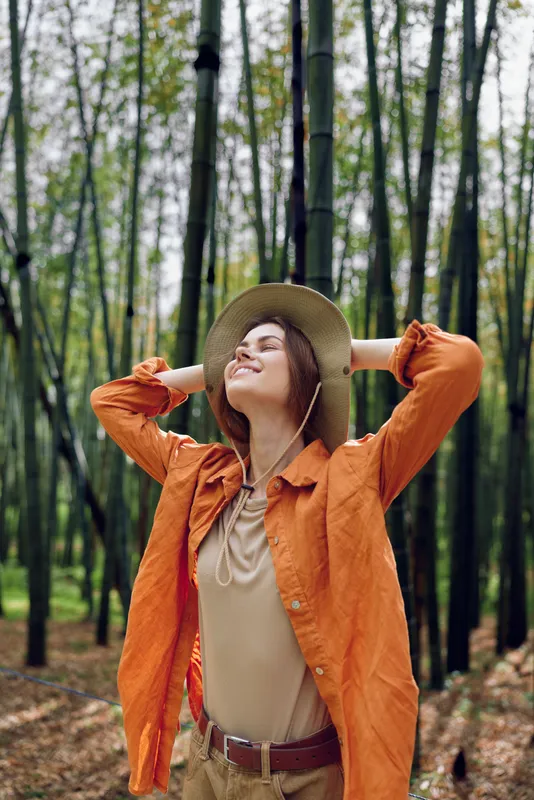 Woman in hat stands in bamboo forest wearing orange jacket, smiling and relaxing with hands behind head, enjoying nature among tall stalks, peaceful green foliage and soft filtered sunlight.