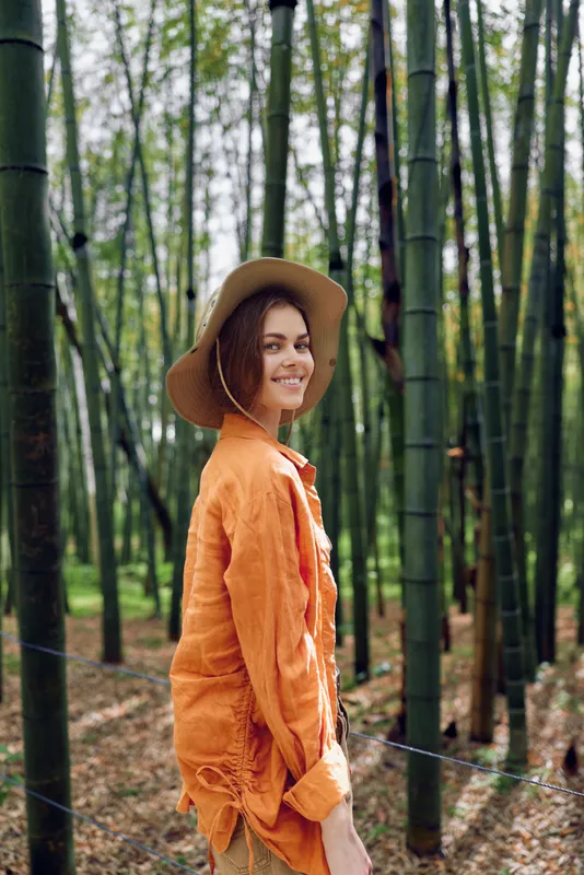 Woman bamboo forest hat smile orange portrait nature travel lifestyle. Young woman in an orange linen shirt and straw hat smiles in a dense bamboo grove during a relaxed outdoor walk.