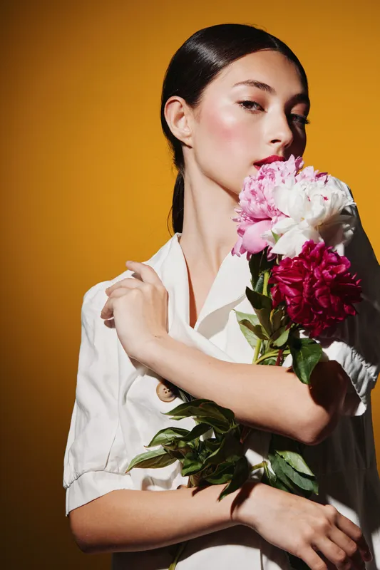 Elegant young woman holds vibrant pink and white peonies embracing them gently. She wears a white blouse with sleek hair against a warm vibrant orange wall. Beauty creative fashion concept.