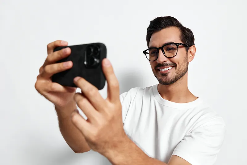Smiling man taking selfie with smartphone, wearing t-shirt and glasses, joyful expression on face, isolated on plain solid background in studio. People lifestyle concept.