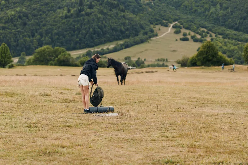 A person in a rural field prepares gear beside a rolled mat, with a horse in the distance. The scene conveys outdoor activity, solitude, and nature exploration.