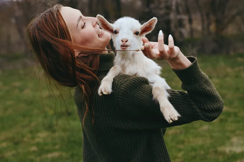 Goat kid and woman share a warm moment outdoors, capturing lifestyle and gentle affection in a grassy countryside setting. Natural light and relaxed pose convey care, happiness, and everyday animal