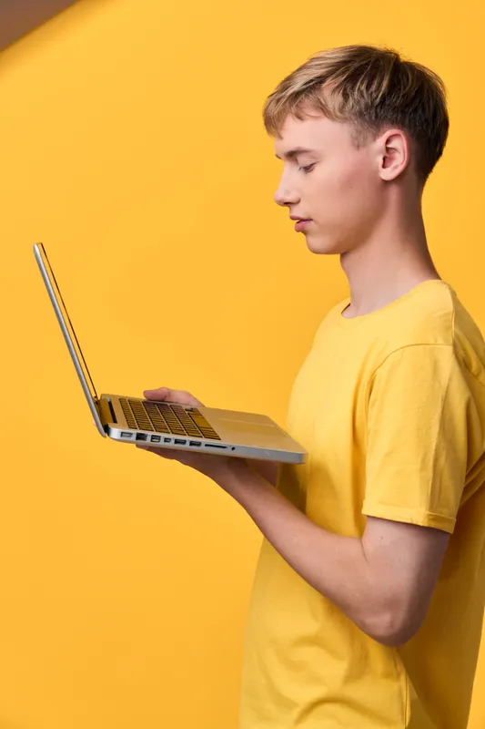 Young man in a yellow t shirt holds a laptop against a bright yellow backdrop, focused on the screen, conveying a modern tech and casual workspace vibe.