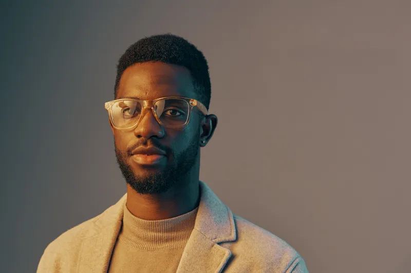 Stylish young Black man in glasses, wearing a beige blazer and sweater, smiling confidently against a soft gradient background ideal for professional portrait use