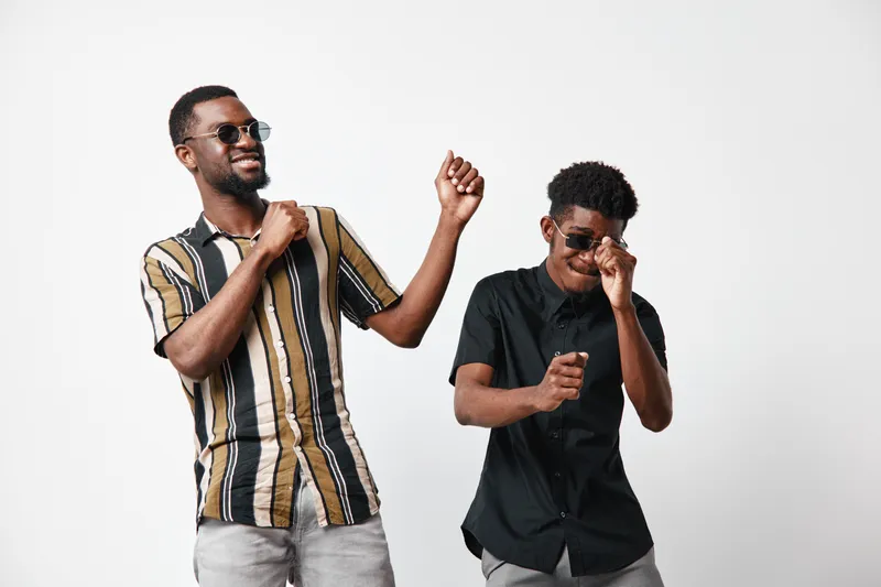 Two black men with dark skin dancing and smiling wearing sunglasses and casual shirts on white background. Friends enjoying music and having fun together in studio.