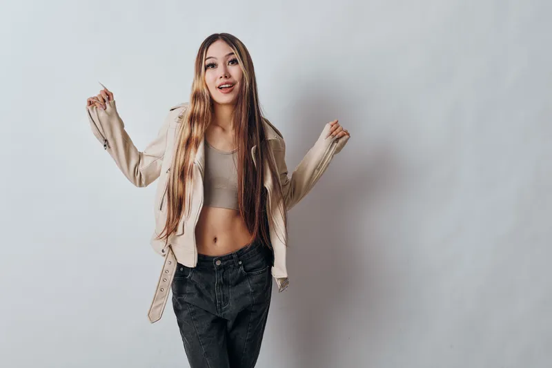 Woman with long hair wearing casual jacket and crop top posing joyfully in studio. Fashion outfit, young adult, smiling expression and confident look on neutral background.