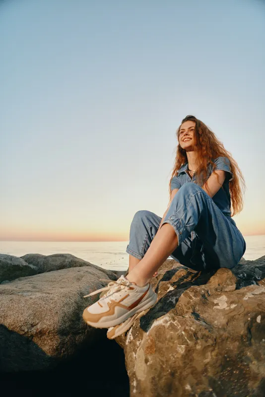 lifestyle woman in modern boho-western denim street style sitting on rocks during golden hour with film color effect, casual sneakers and relaxed smile outdoors