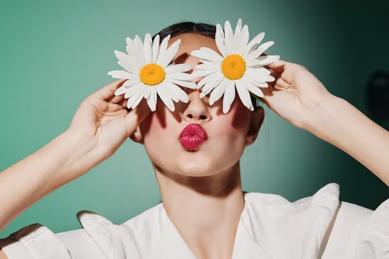 Young woman with glossy pink lips holds two large white flowers over her eyes. She puckers her lips in a playful kiss. Creative beauty fashion portrait on teal background.