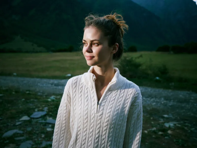 Woman wearing a white cable knit sweater stands in a mountain meadow, calm and thoughtful, amid scenic landscape and soft natural light for a serene outdoor moment