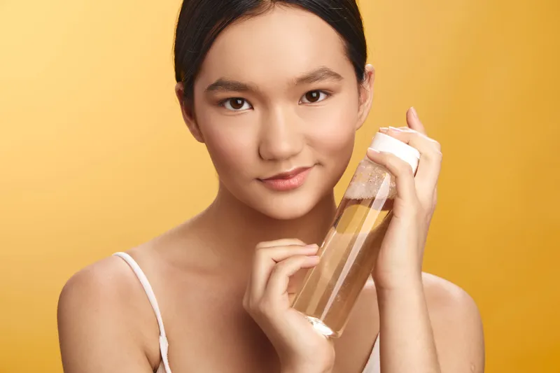 Asian woman holds a clear bottle near her face, presenting a skincare moment with bright studio lighting, a calm smile, and a sense of fresh beauty and selfcare.