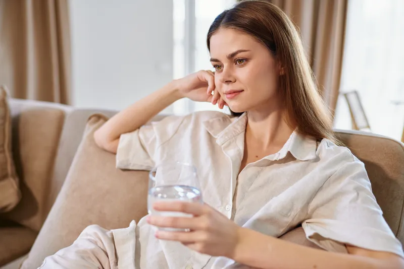 Woman on sofa holding glass of water at home, relaxed and thoughtful portrait in casual loungewear, sitting comfortably in livingroom enjoying calm hydration and quiet moment.