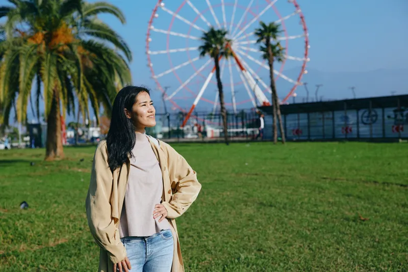 A woman in a beige coat stands in a sunny park with palm trees and a large ferris wheel in the background, showcasing casual fashion and outdoor leisure in a bright setting