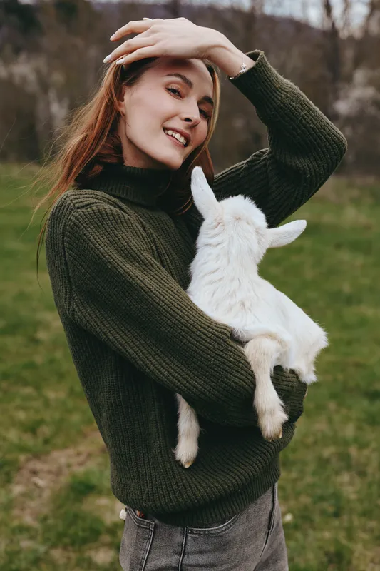 woman goat kid outdoors lifestyle joyful smiling sweater field countryside sunlit nature affection cuddle animal companion rural grass meadow, a warm, candid moment captured with natural colors and
