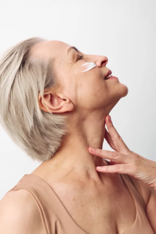 Mature woman with short grey hair applies face cream on cheek, enjoying skincare routine. Concept of natural beauty, aging gracefully, self-care, and wellness in studio lighting.