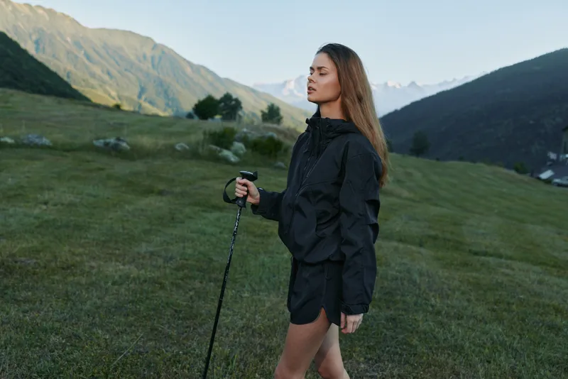 A woman hiking in a rugged mountain landscape, dressed in a black rain jacket and shorts, holding a trekking pole as she gazes into the distance with determination.